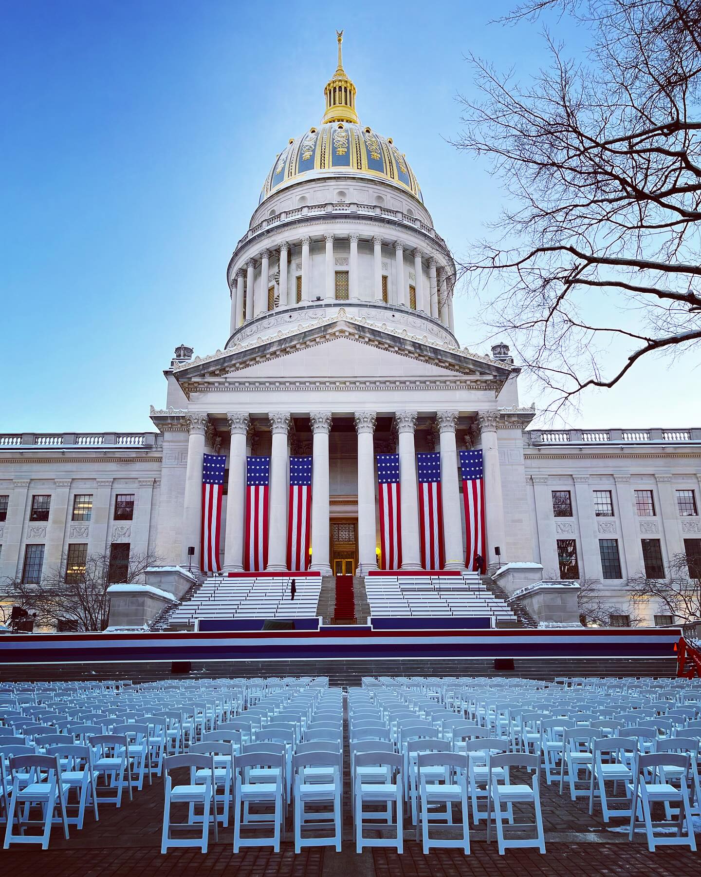 WV State Capitol with gold dome, US flags, and rows of white chairs on a snowy day for an inauguration.