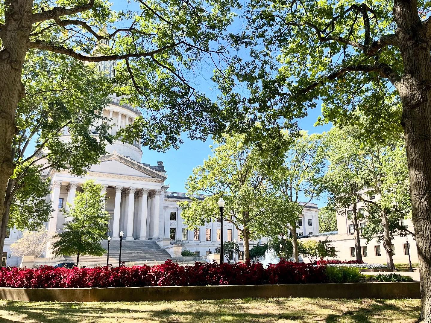 WV State Capitol dome framed by lush green trees and red flowers under a blue sky.