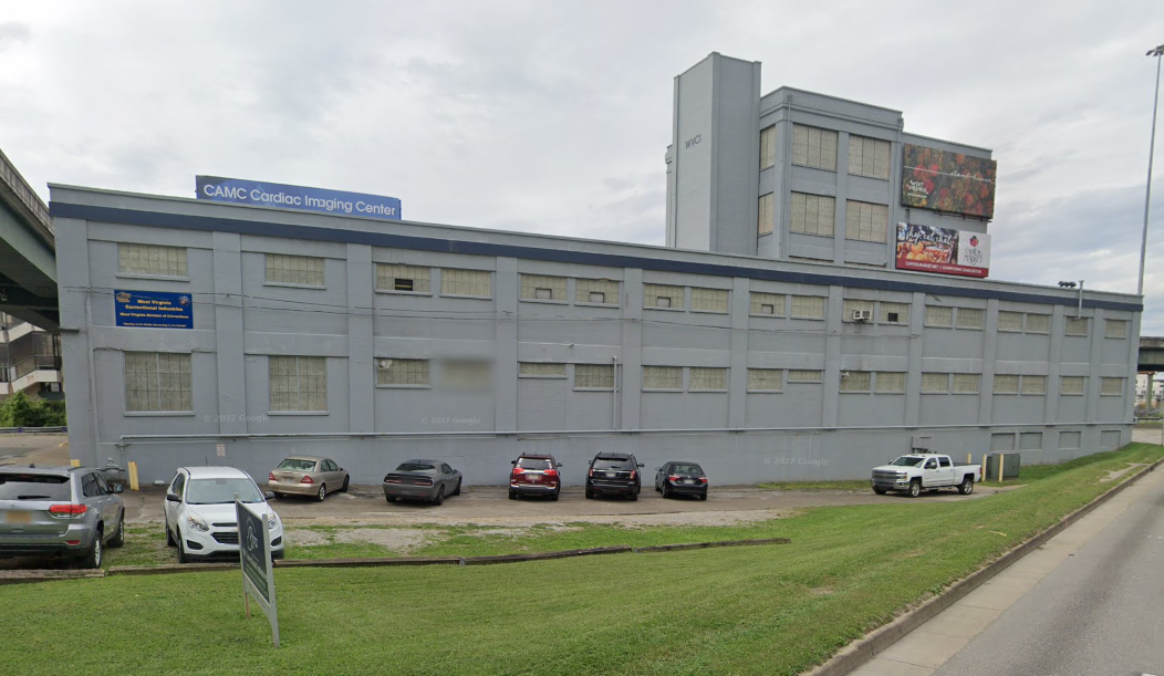 Large gray industrial building with rows of small windows, a taller rear section, and cars parked on a narrow gravel lot.